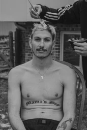 Shirtless man sitting on a chair while someone applies hair dye with a brush, captured in black and white by Emily Maye.