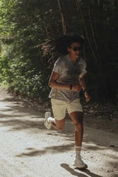A man with long dreadlocks running on a dirt path through a dense forest wearing a grey t-shirt and light shorts.