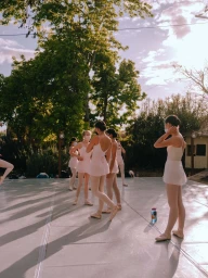 Ballet dancers in pink leotards and tutus rehearsing outdoors on a stage during late afternoon sunlight.