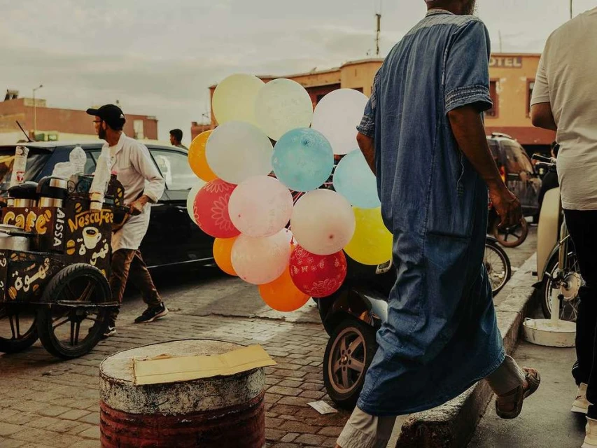 Outdoor scene with an older adult walking past colorful balloons on a scooter, a vendor with a coffee cart, and architecture in the background.