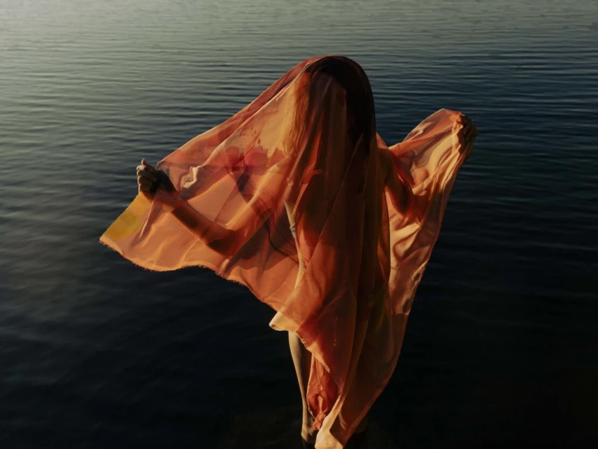 Person stands in water, partially covered by orange fabric that obscures their face, illuminated by golden sunlight at dusk.