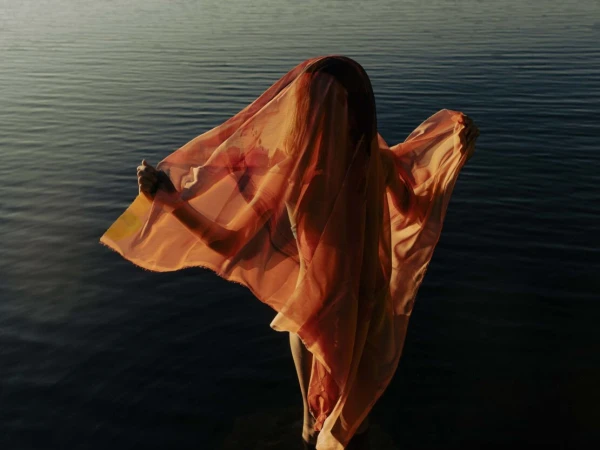 Person stands in water, partially covered by orange fabric that obscures their face, illuminated by golden sunlight at dusk.