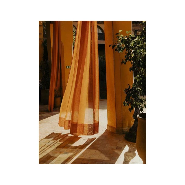 Sheer orange curtains hanging between yellow columns, creating patterns of light and shadow on the tiled floor of an outdoor space.