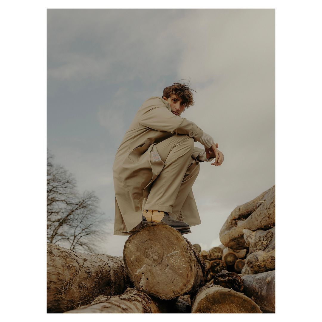Man in beige trench coat squats on logs outside with overcast sky in fashion editorial, portrait orientation, eye-level angle.