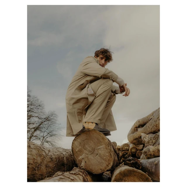 Man in beige trench coat squats on logs outside with overcast sky in fashion editorial, portrait orientation, eye-level angle.