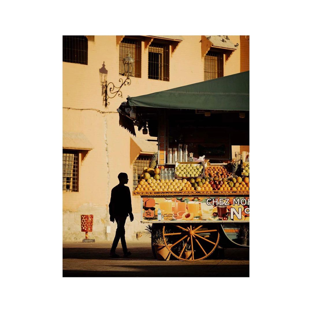 Silhouette of a man walking past a fruit and juice stand in an urban environment with yellow buildings in the background.