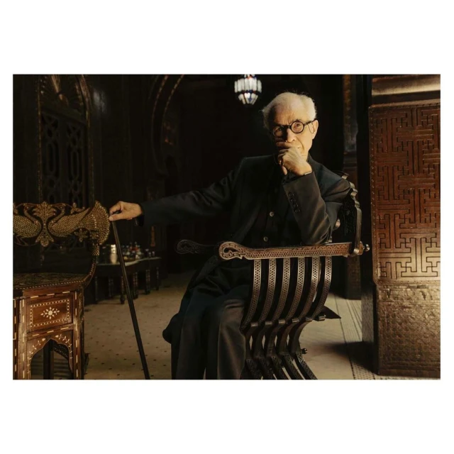 Elderly man in glasses seated in an ornate chair with hand to face in a dark, elaborately decorated indoor environment.