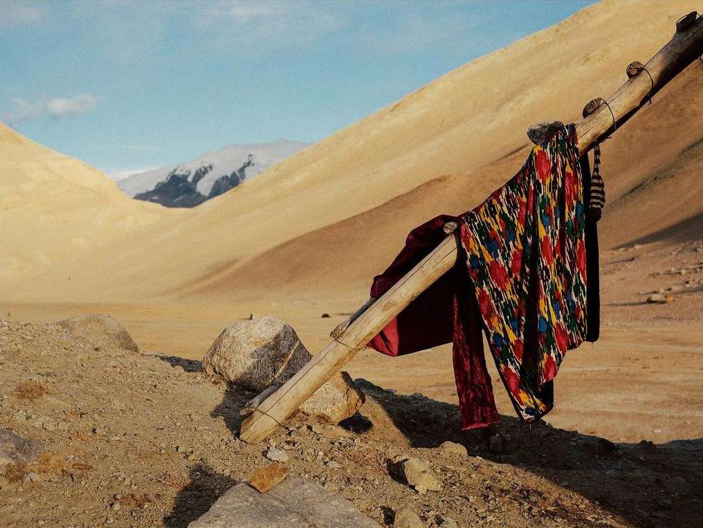 Traditional Uzbek ikat textile draped over a weathered wooden frame, set against a backdrop of arid mountains under a clear sky.