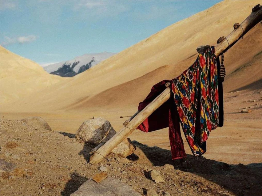 Traditional Uzbek ikat textile draped over a weathered wooden frame, set against a backdrop of arid mountains under a clear sky.