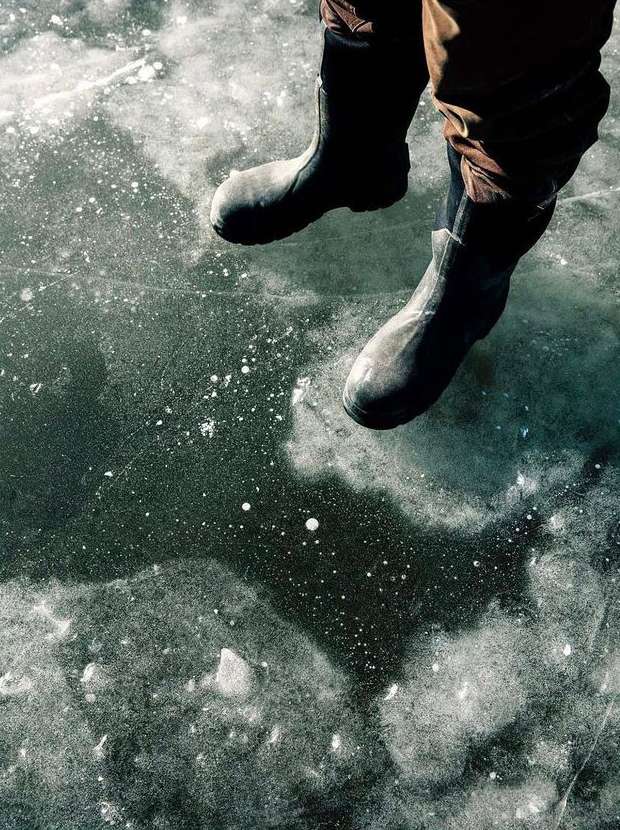 Close-up shot of a person wearing black rubber boots while sitting on a frozen body of water, the ice reflecting the surrounding light.