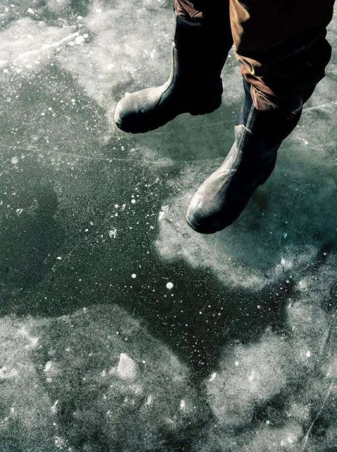 Close-up shot of a person wearing black rubber boots while sitting on a frozen body of water, the ice reflecting the surrounding light.