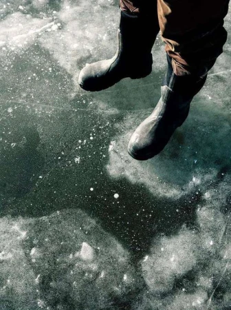 Close-up shot of a person wearing black rubber boots while sitting on a frozen body of water, the ice reflecting the surrounding light.