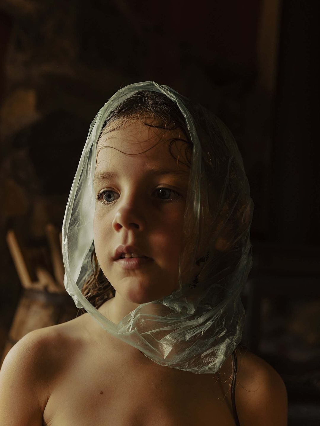 Portrait of a child with a clear plastic bag on their head, looking off camera in a dimly lit indoor environment.