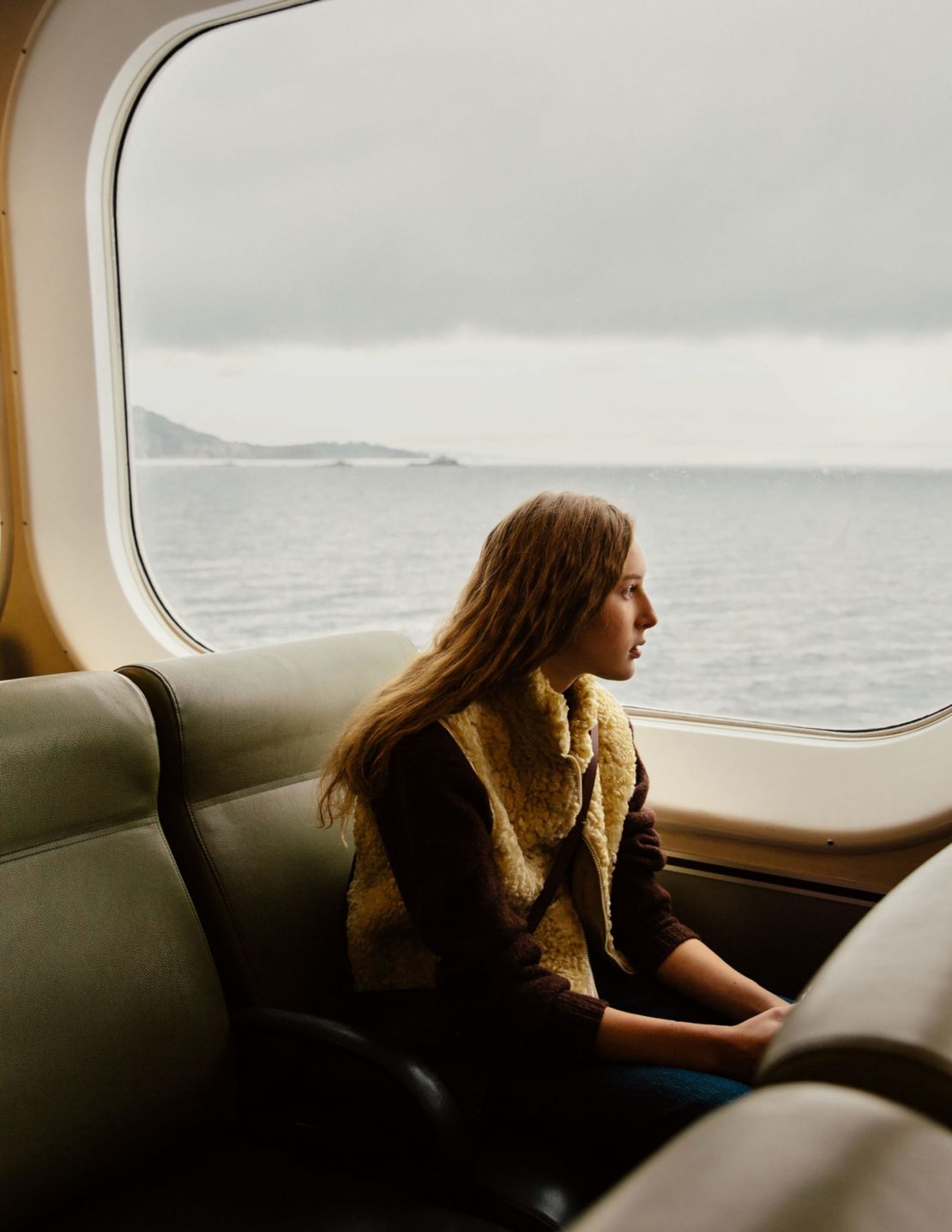 Woman in a vest sitting inside a ferry looking out the window at the ocean on a cloudy day with a coastal view in the background.