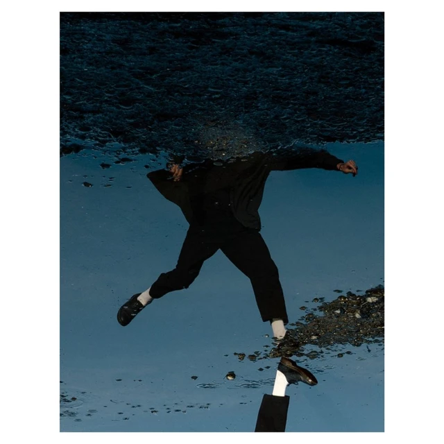 Reflection of a person walking in water, wearing dark clothes, with a second person's arm and shoe visible, creating a surreal effect.