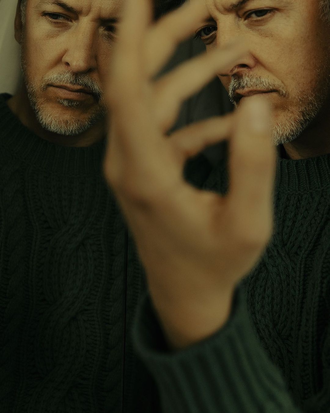 Close-up of an adult man with salt and pepper hair and beard wearing a green sweater, reflected in a mirror with blurred hand.