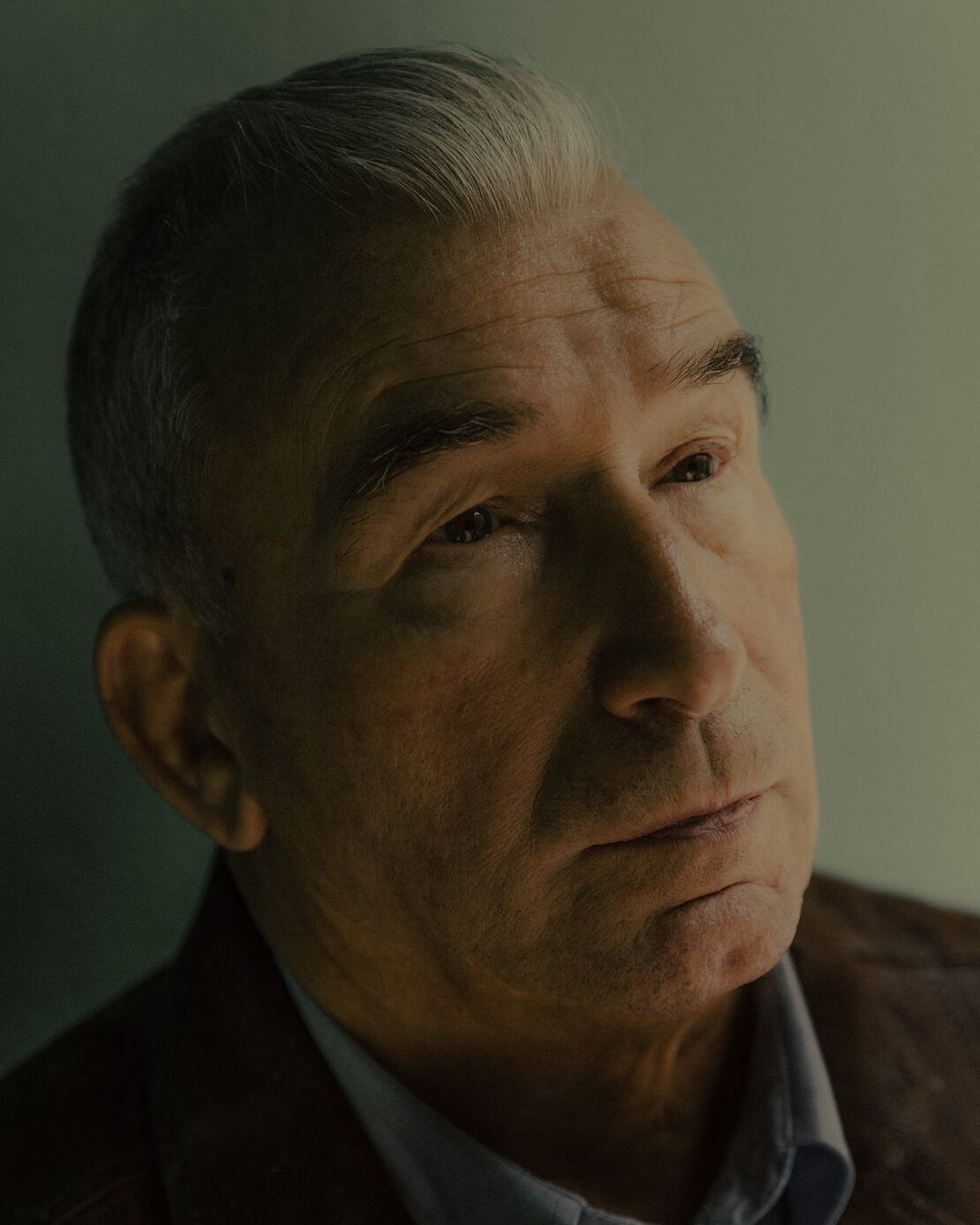 Portrait of mature adult man with grey hair wearing a collared shirt and jacket in an indoor setting with dramatic lighting.