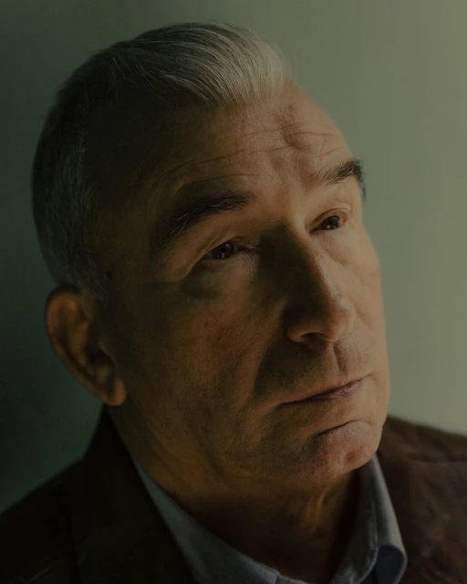 Portrait of mature adult man with grey hair wearing a collared shirt and jacket in an indoor setting with dramatic lighting.