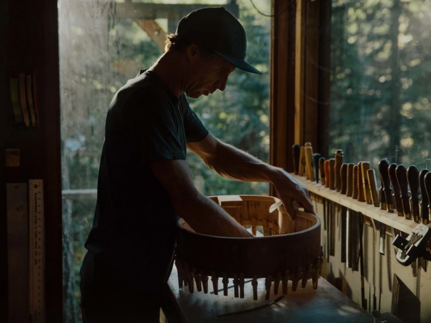 Adult male craftsman working in a woodworking studio, shaping a round wooden frame near an open window and assorted woodworking tools.