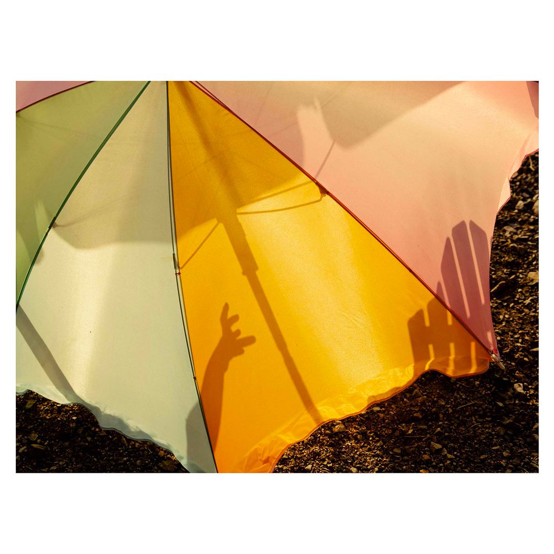 Sunlit multicolored umbrella casts shadow of a hand. Segments of yellow, green, and pink form a vibrant canopy, casting playful shadows.