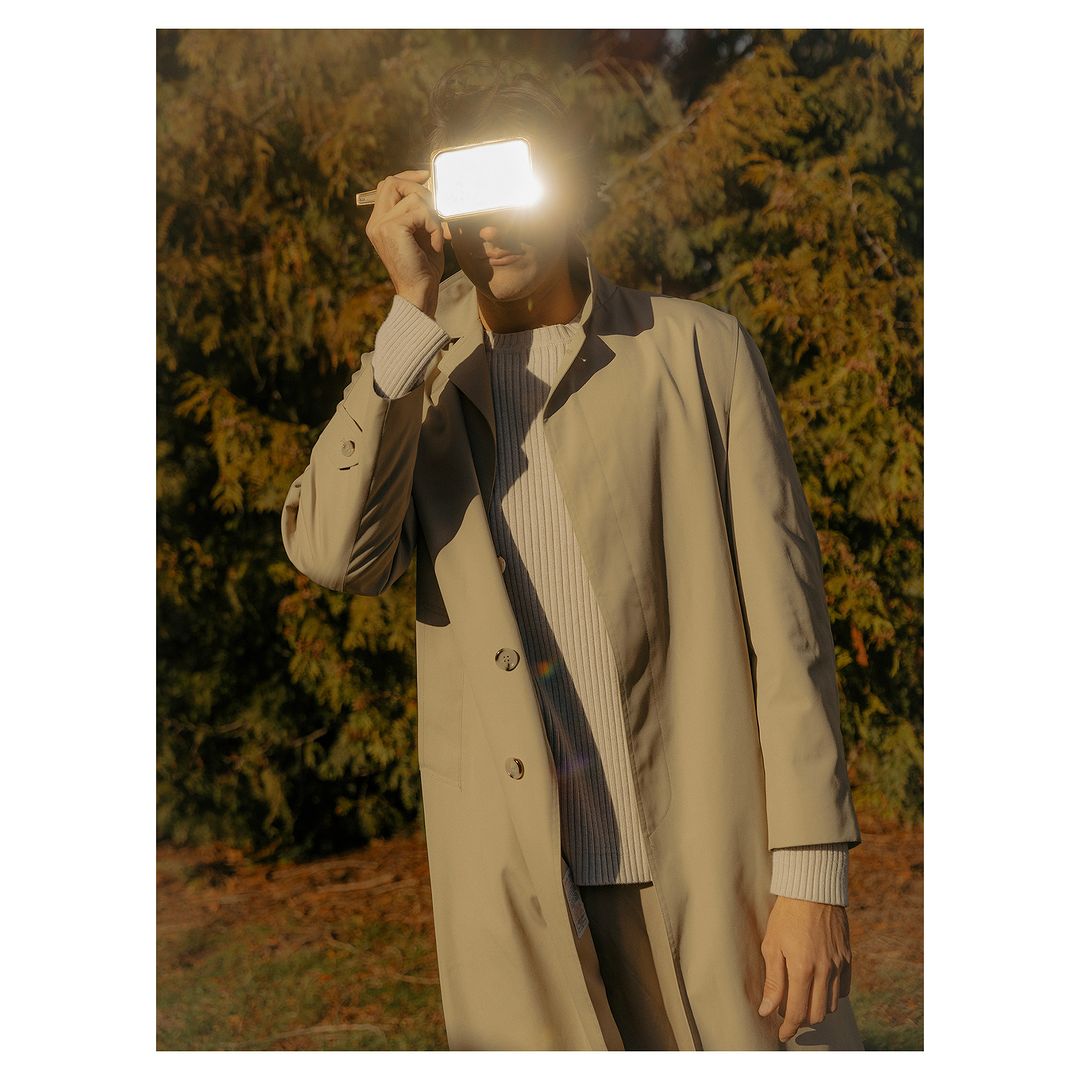 An adult male in a trench coat poses outdoors, holding a bright light panel in front of his face, with blurred trees in the background.