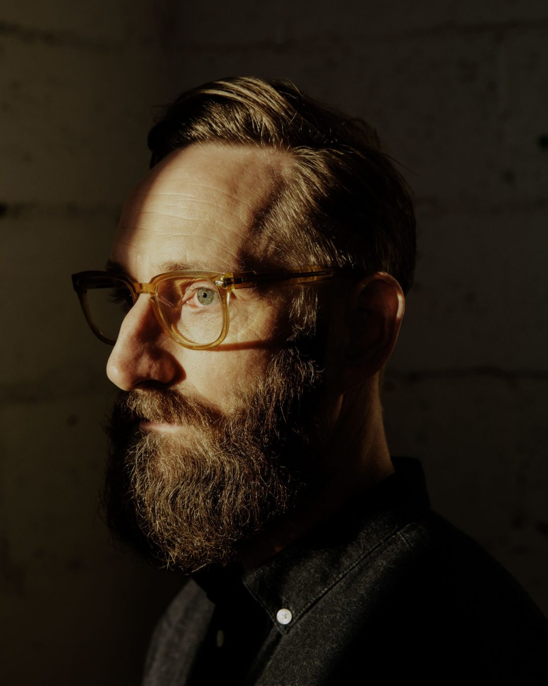 Profile portrait of a bearded man wearing clear-framed glasses and a dark button-down shirt.