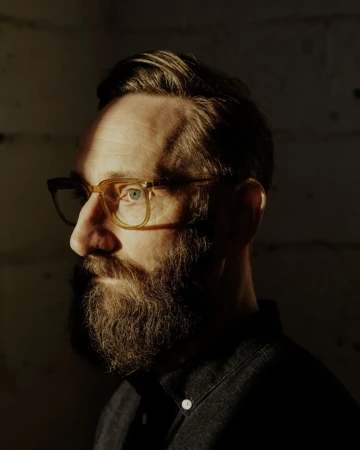 Profile portrait of a bearded man wearing clear-framed glasses and a dark button-down shirt.