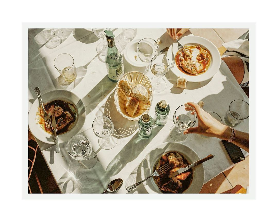 Overhead view of a sunlit table with several diners sharing plates of pasta, wine bottles, and glasses during an outdoor meal.