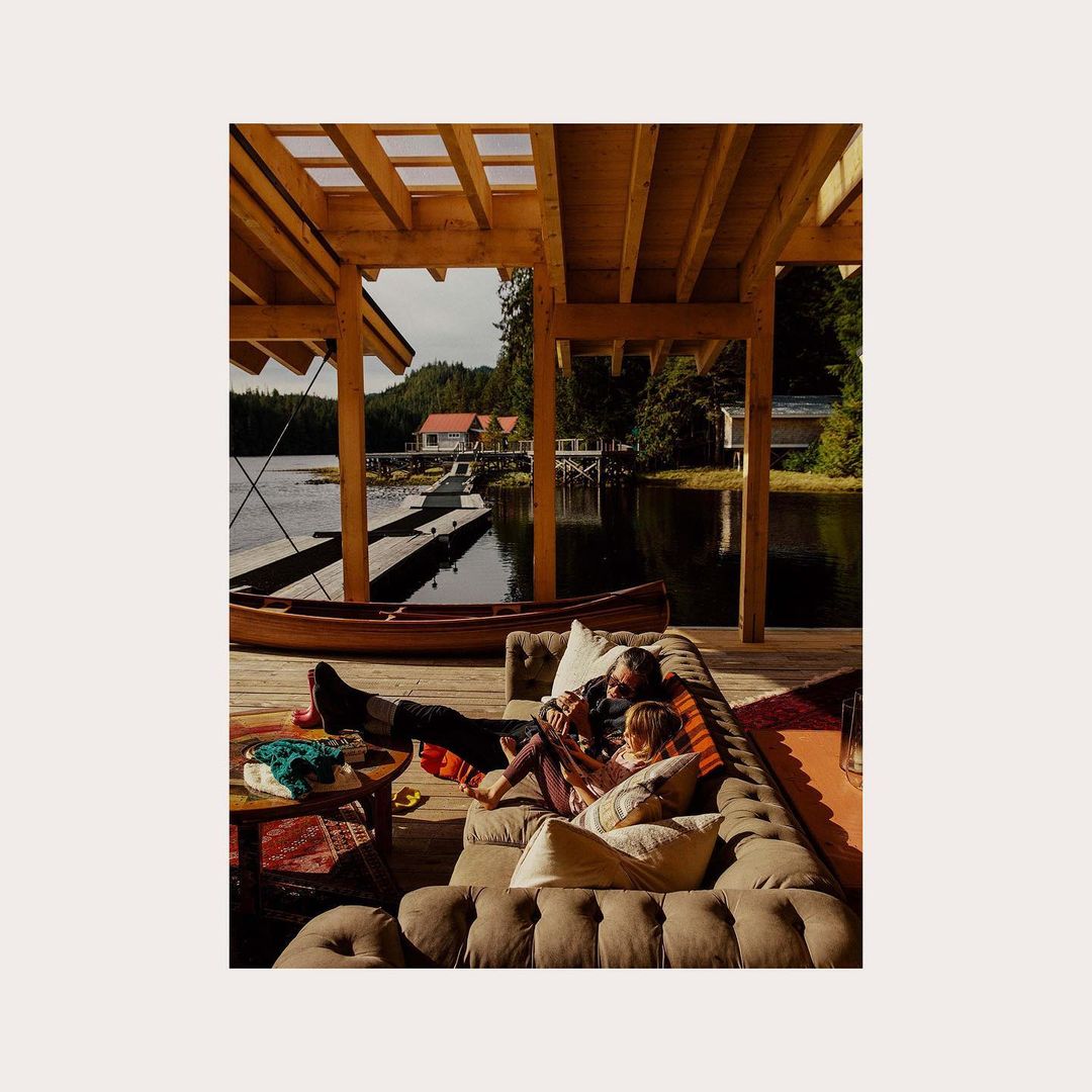 Father and child relax together on an outdoor couch at a lakeside cabin; dock, boat, and trees in the background; sunny daylight.