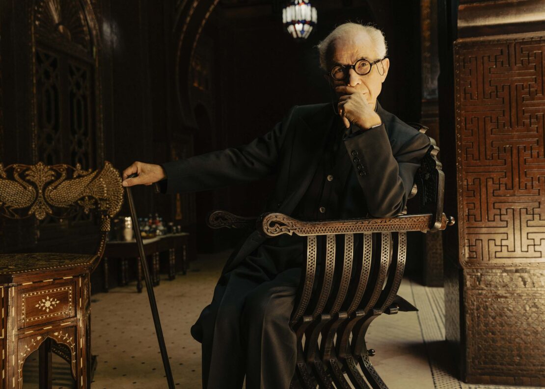Elderly man in a dark suit sits in an ornate chair holding a cane in an elaborate room with patterned wood panels.