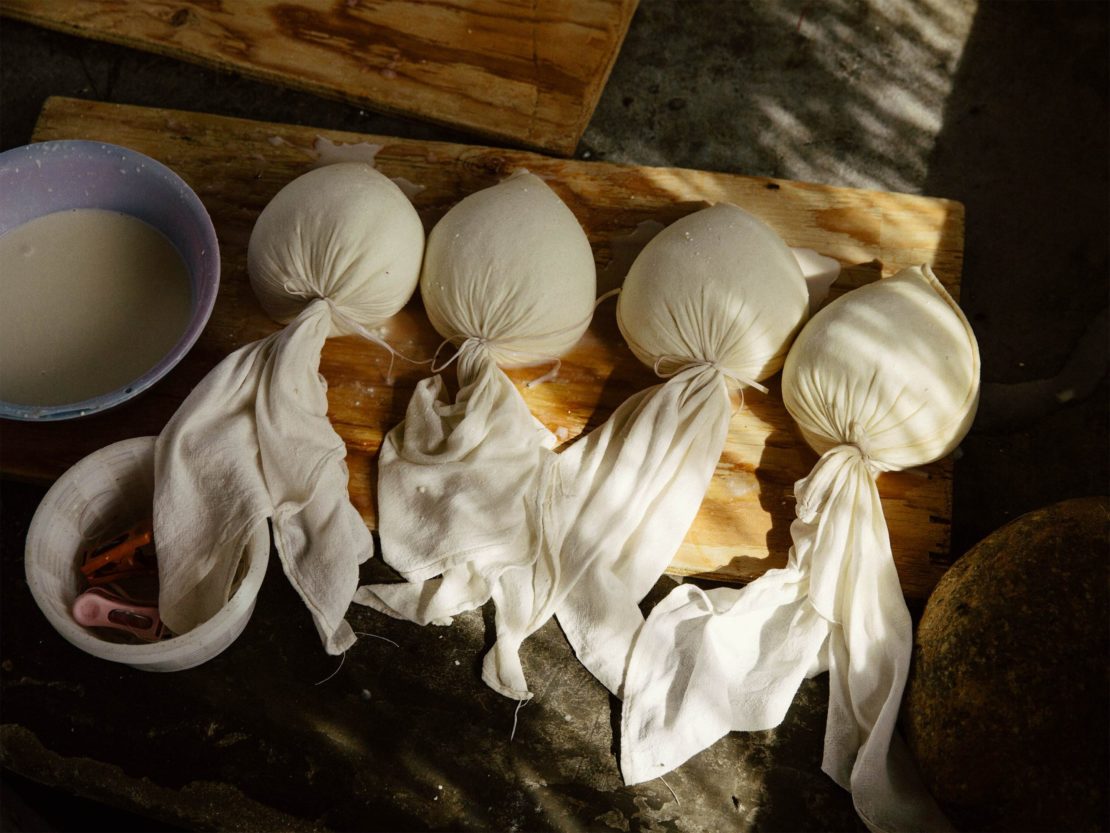 Overhead shot of four cheesecloth sacks containing white cheese curds on a wooden surface, with bowls holding liquid and utensils.