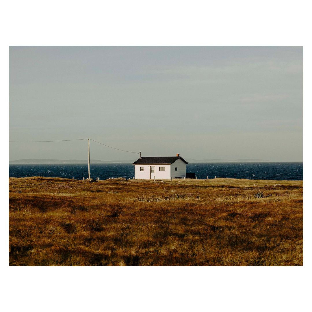 A single-story white house with a black roof sits in a grassy field by the ocean under an overcast sky, creating a serene and remote scene.