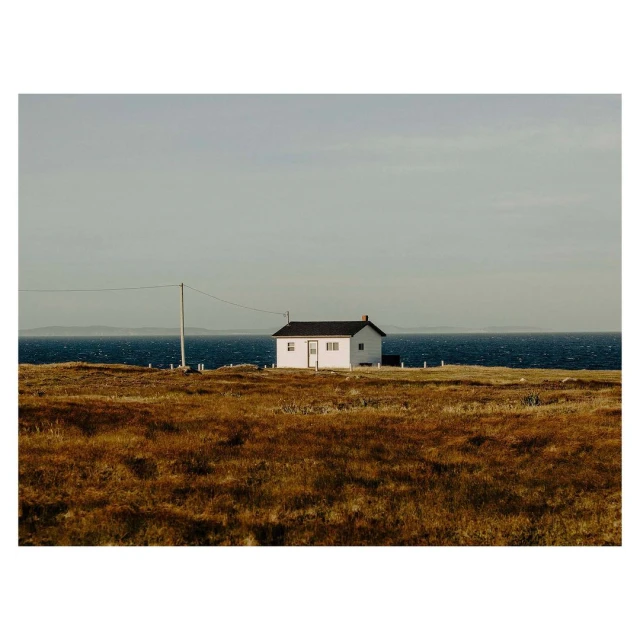 A single-story white house with a black roof sits in a grassy field by the ocean under an overcast sky, creating a serene and remote scene.