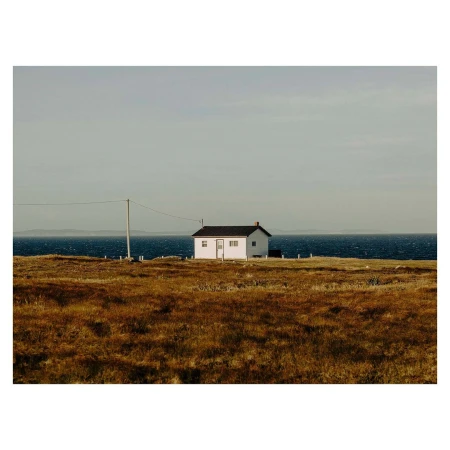 A single-story white house with a black roof sits in a grassy field by the ocean under an overcast sky, creating a serene and remote scene.