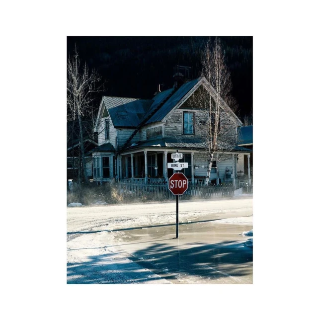 An old weathered house stands beside a stop sign on a street covered in water or ice. Dark mountains are visible in background.