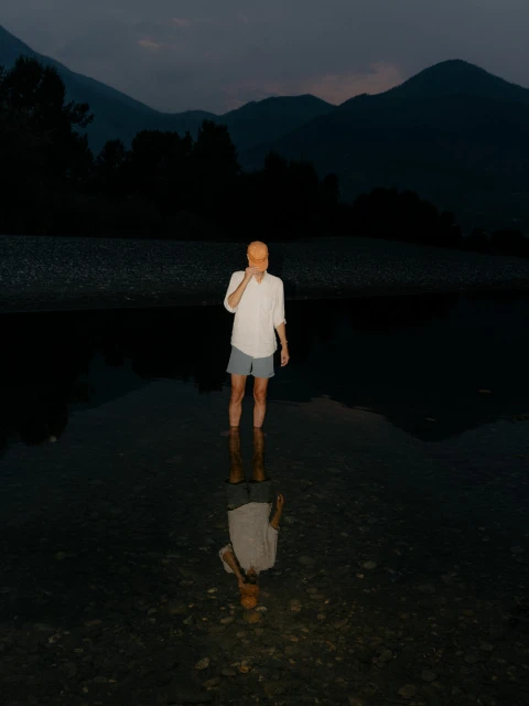 A man in shorts and a shirt stands in a shallow river with mountains in the background. The image reflects in the dark water.