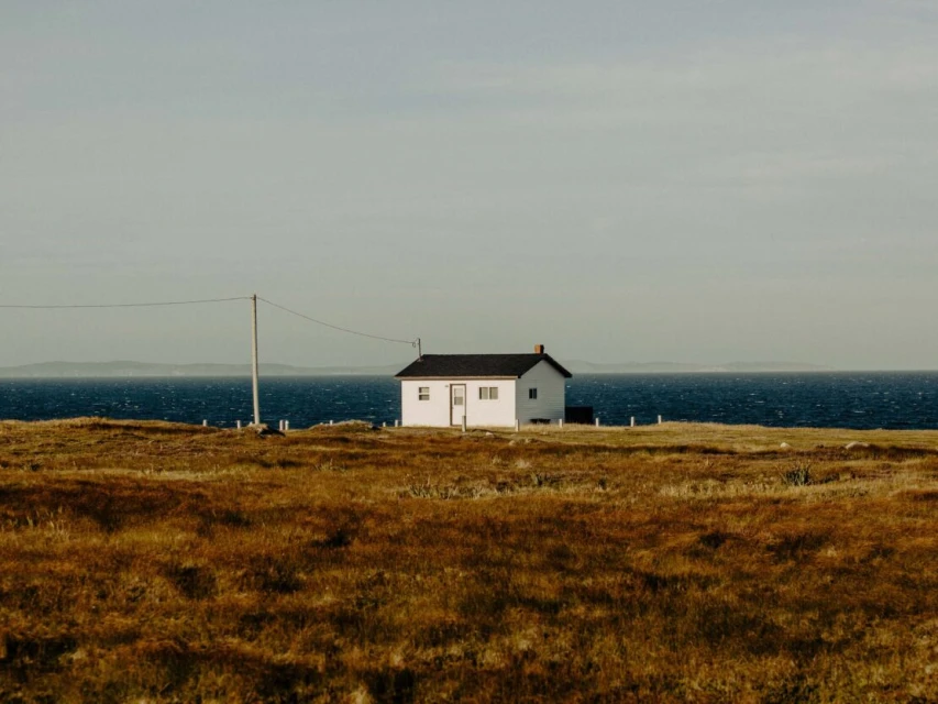 White house with black roof on grass field near ocean and horizon under bright cloudy sky, power line visible, landscape format.