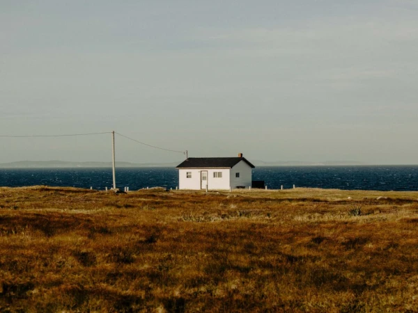 White house with black roof on grass field near ocean and horizon under bright cloudy sky, power line visible, landscape format.