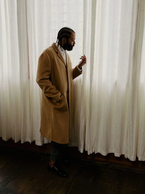 Portrait of a stylish adult male wearing a tan coat standing by a window with sheer white curtains, looking off to the side.