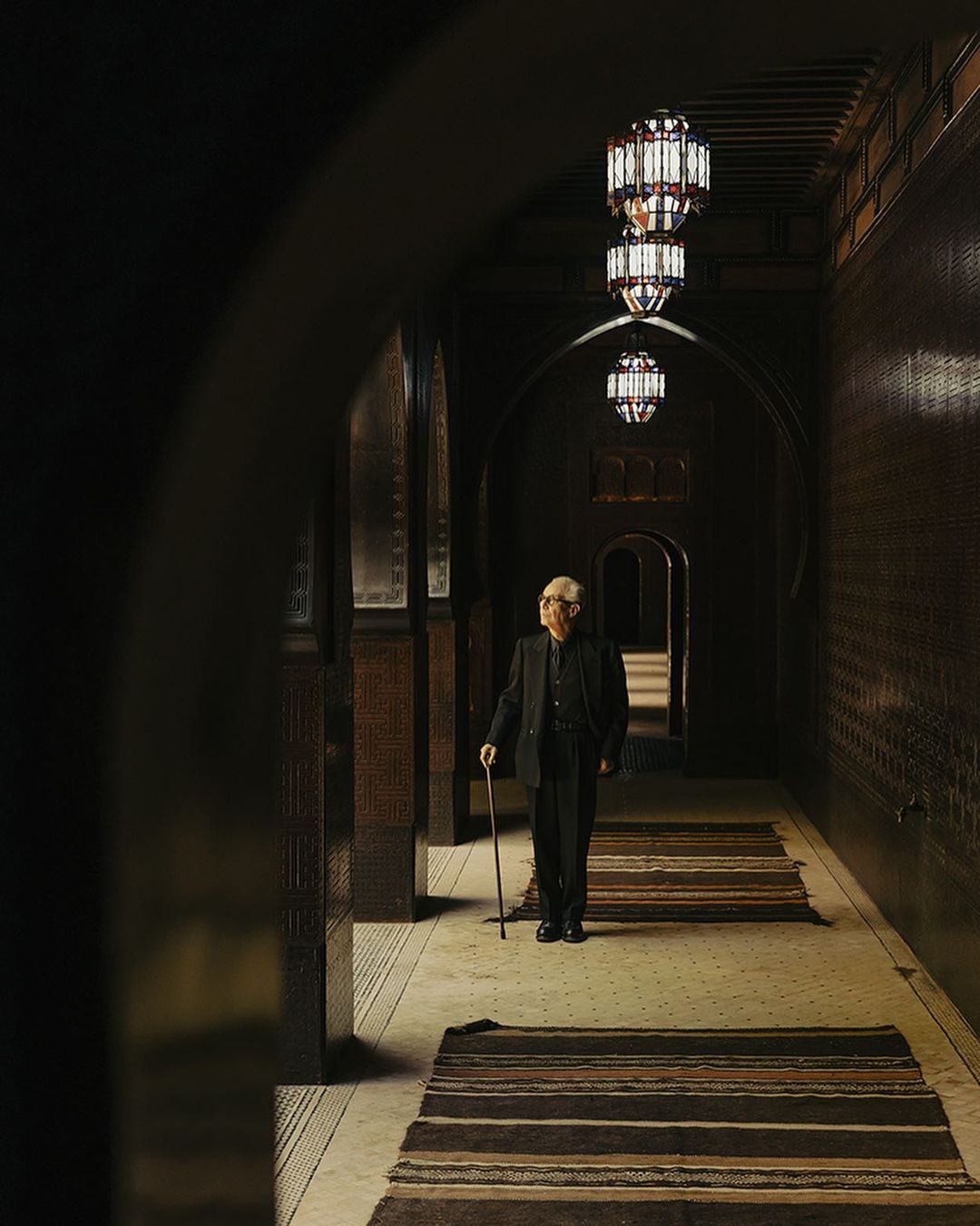 Elderly man in a suit walking in a hallway with decorative lights and carpets. Portrait of elegance and history.