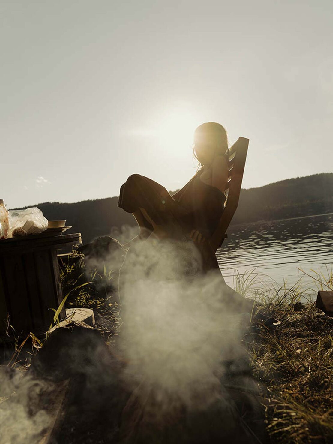 Silhouette of a person sitting in a chair by a lake at sunset with smoke rising from a campfire in the foreground.