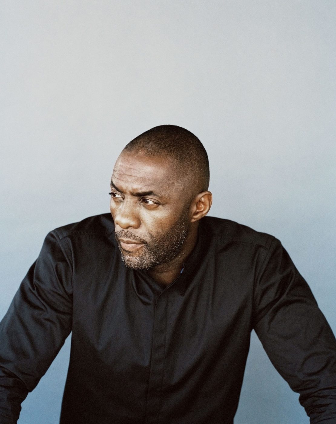 Portrait of Idris Elba in a black shirt, looking off-camera against a grey backdrop. Fashion and celebrity lifestyle photography.