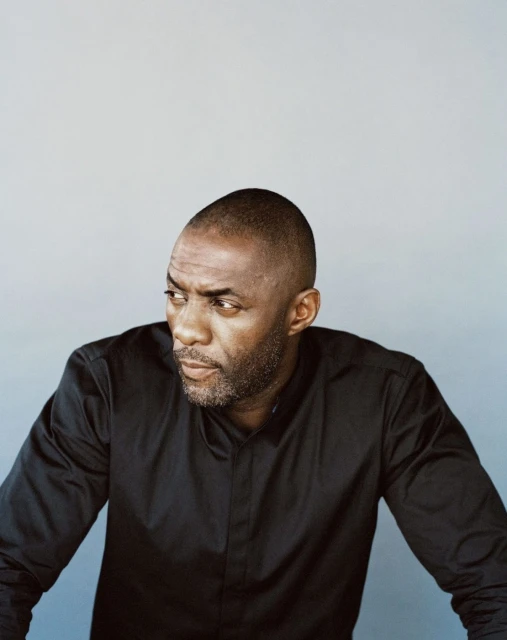 Portrait of Idris Elba in a black shirt, looking off-camera against a grey backdrop. Fashion and celebrity lifestyle photography.