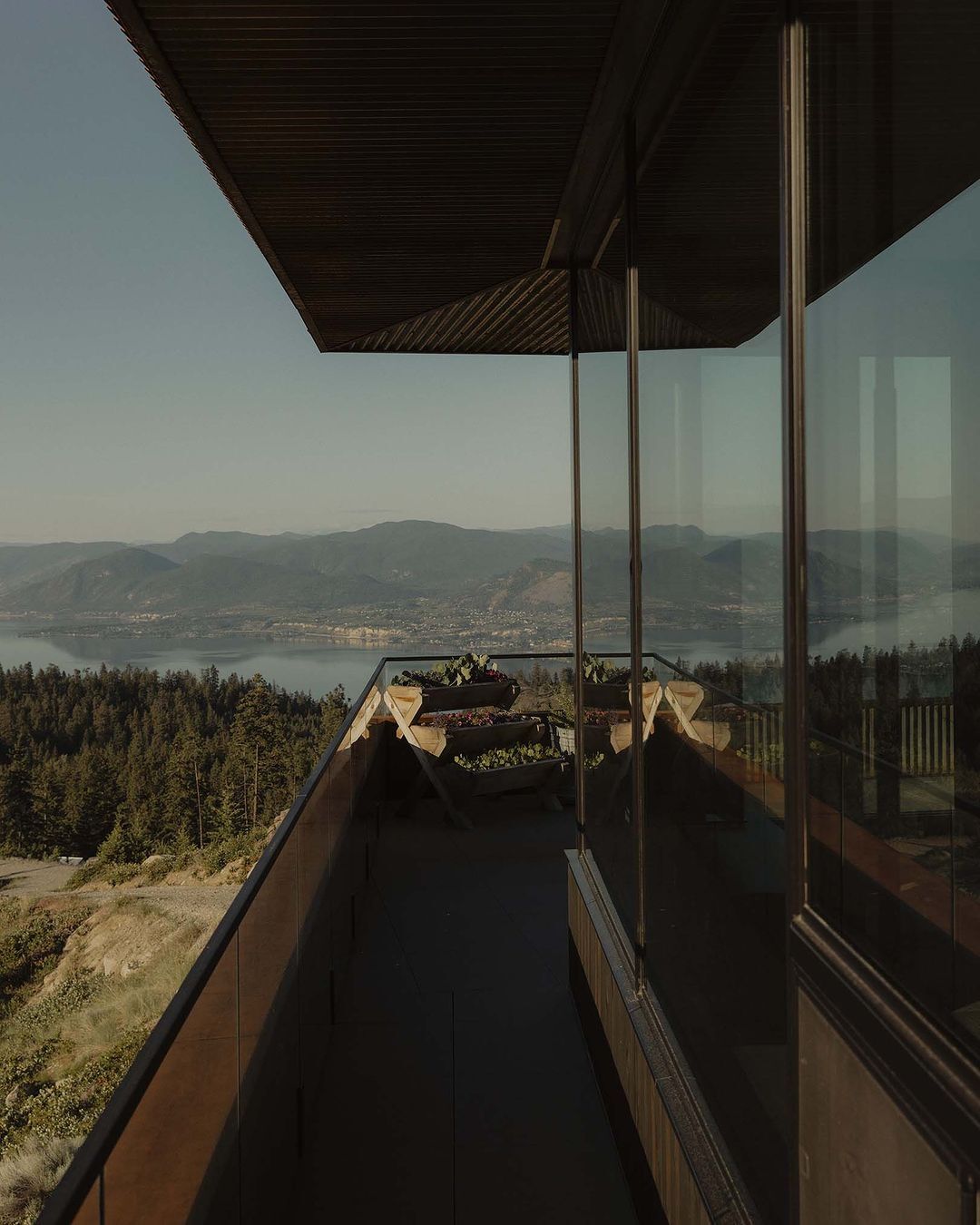 Modern architecture featuring a glass-walled balcony overlooking a mountain and lake landscape, bathed in soft daylight.