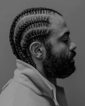 Profile portrait of a man with cornrows and a beard wearing a high-collared jacket against a grey background.