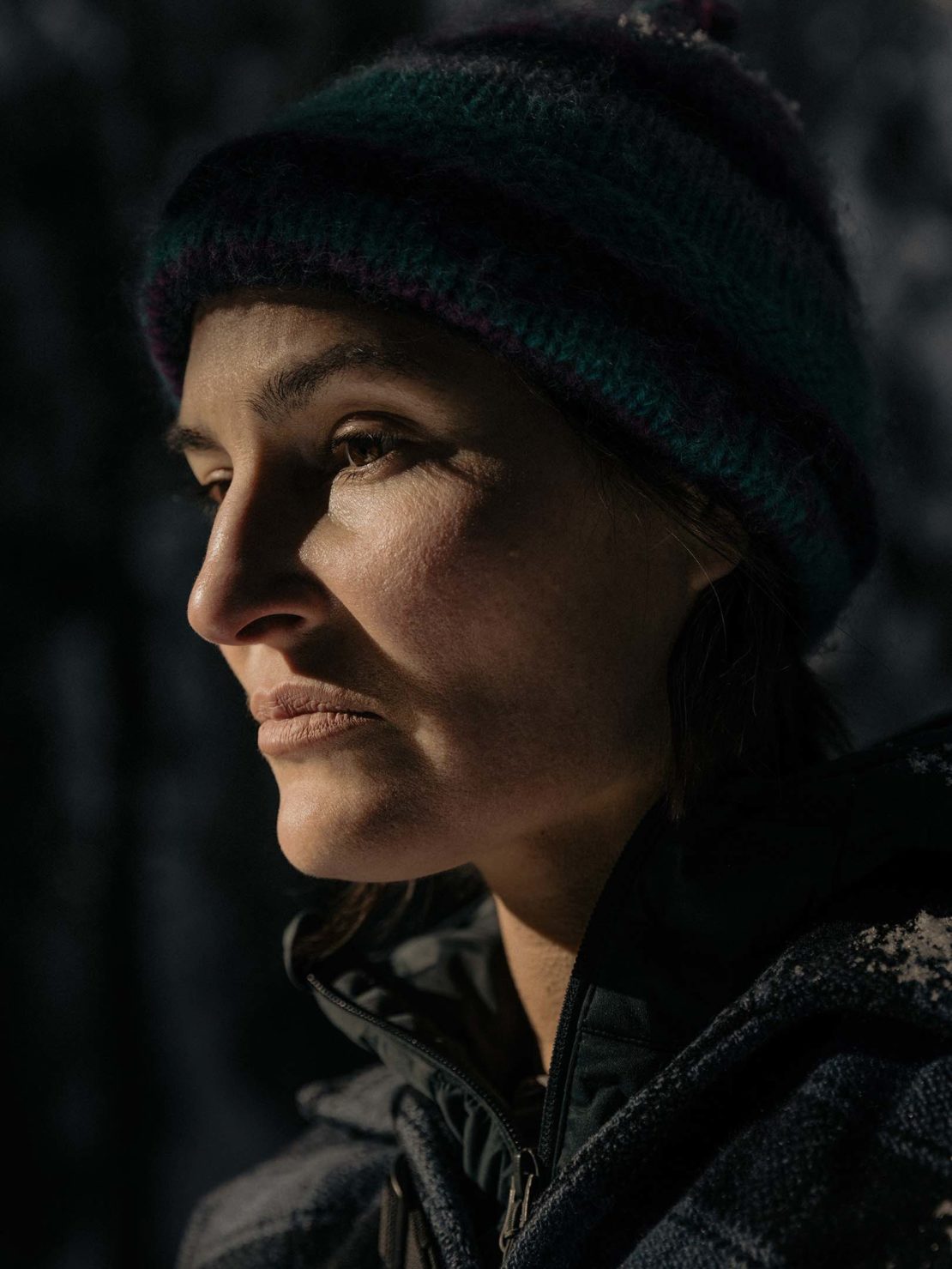 Close-up of a woman in a wool hat looking left. She is wearing a dark jacket with a gray, patterned fleece and is outside in soft light.