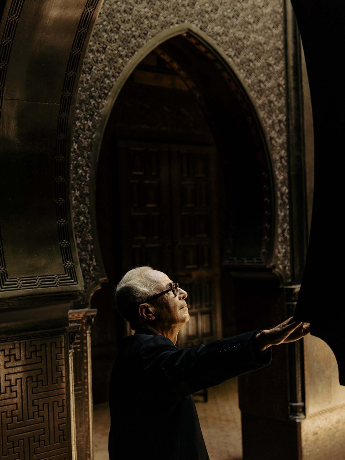 Elderly man in dark suit stands beneath ornate Moorish archway, eyes upward, hand extended, captured in soft, ambient lighting.