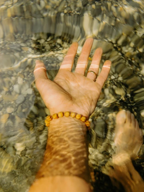 An arm and leg emerge from clear water with pebble reflections, showing a beaded bracelet and a gold ring on a finger.