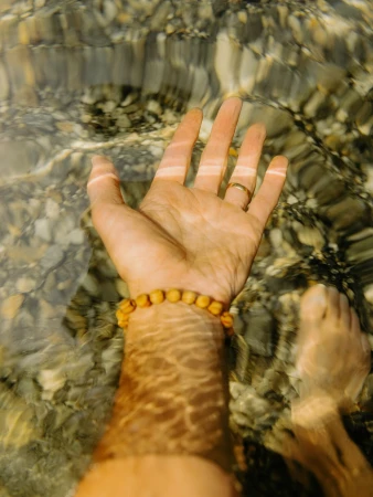 An arm and leg emerge from clear water with pebble reflections, showing a beaded bracelet and a gold ring on a finger.