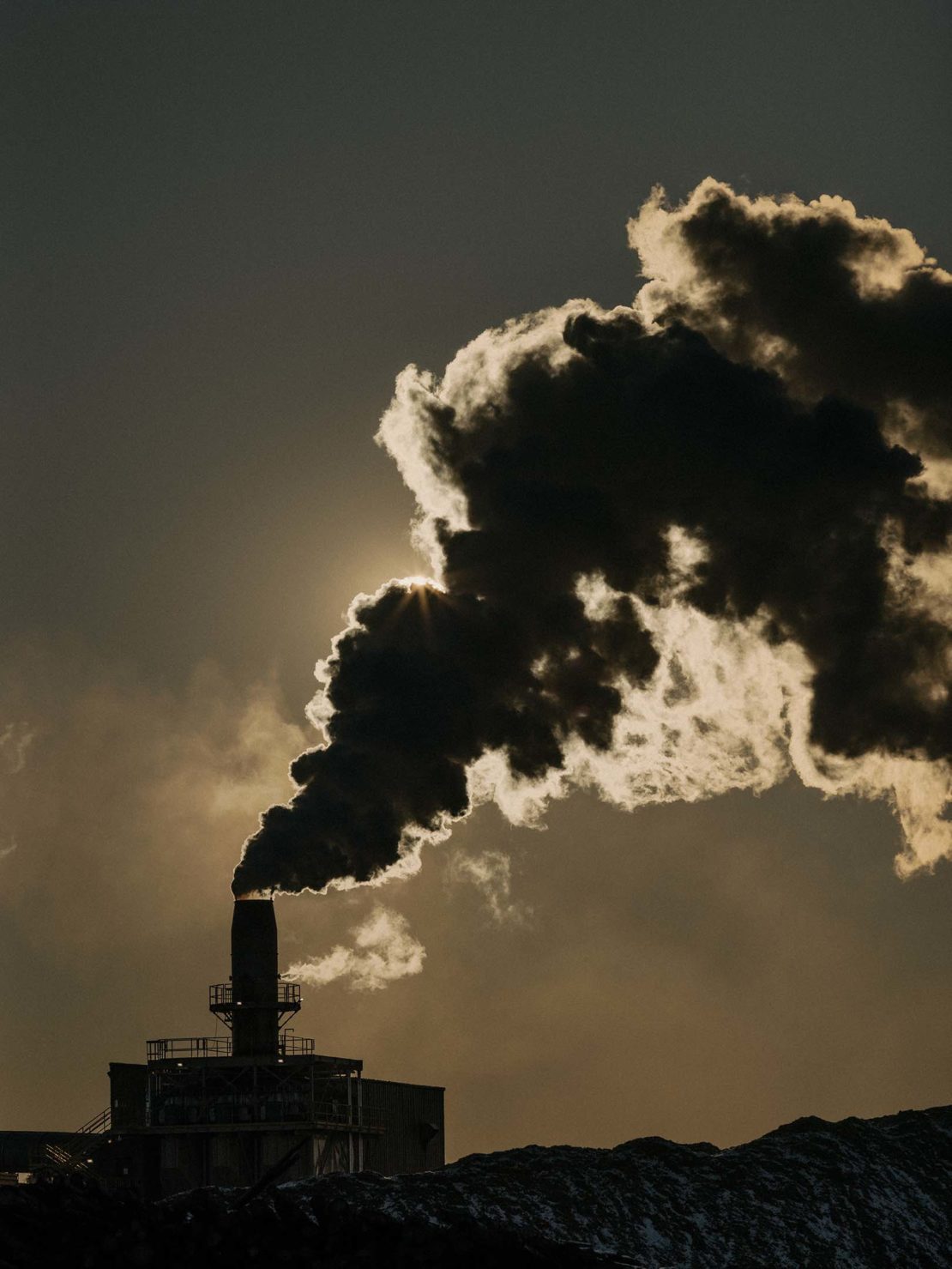 Industrial plant with smokestack emitting dark smoke against a cloudy sky with sun rays, symbolizing industry and potential environmental concerns.